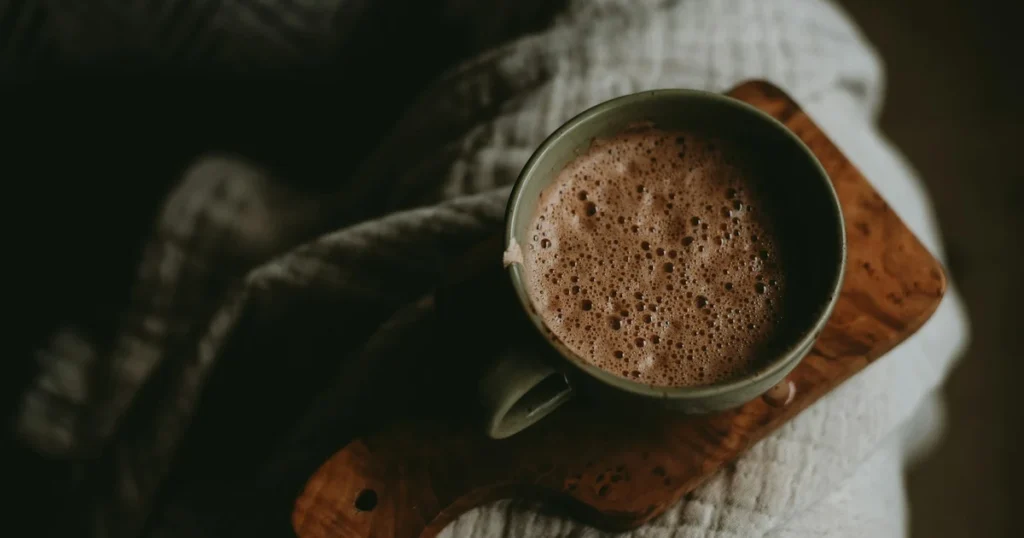 Hands holding a steaming mug of dark ceremonial cacao with soft window light illuminating the rising steam